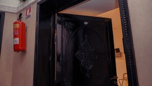 A black ornate door with a number '5' above it, a fire extinguisher mounted on the wall to the left, and a chair in the background. - Riad Roummana Marrakech (Marrakech)