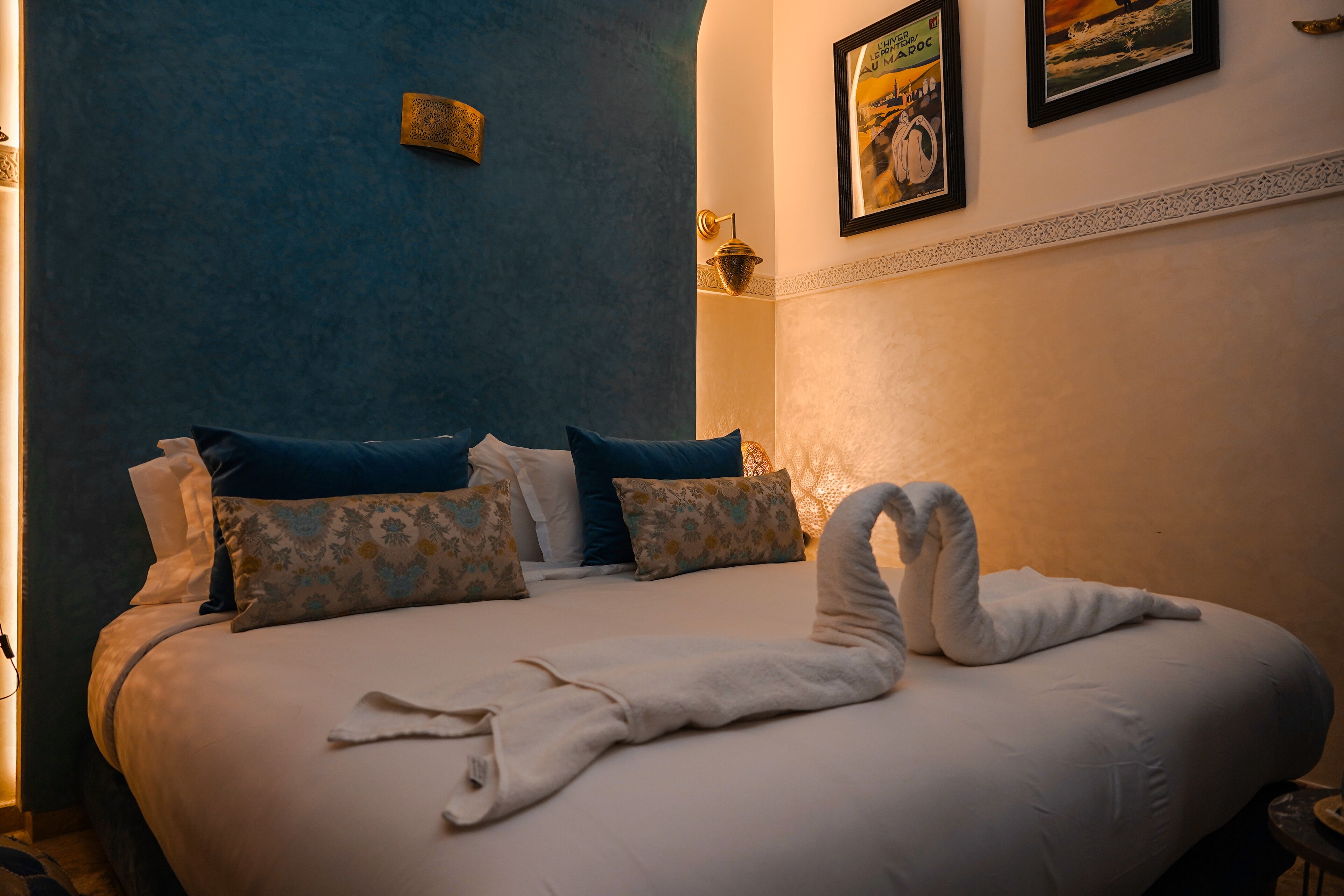 A neatly made bed with a white blanket and a towel arranged in a swan shape, blue and patterned pillows, and a wall with a framed picture and a decorative light fixture.