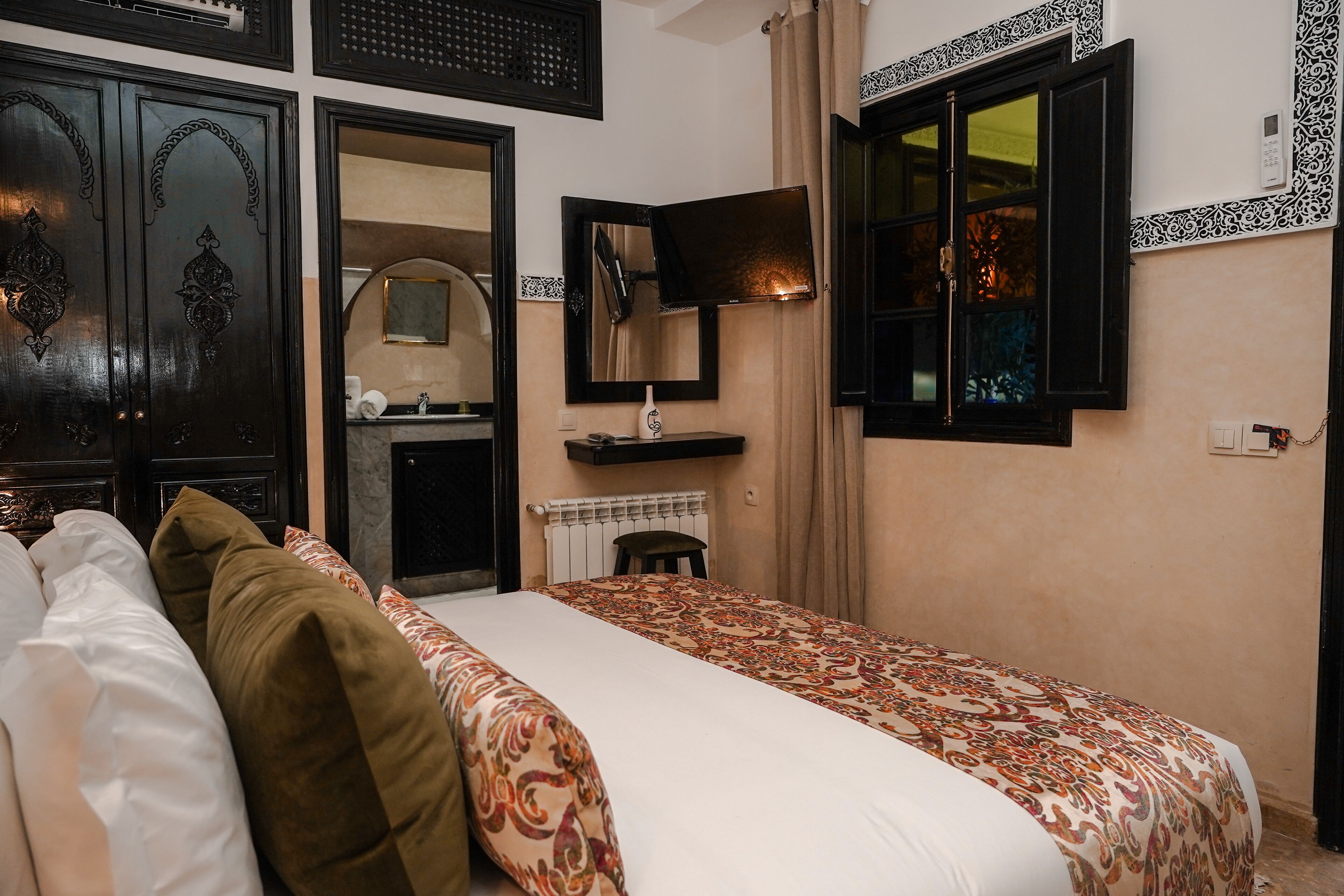 A hotel room with a bed, a bedside table, a mirror, a window with curtains, and a wall-mounted TV.