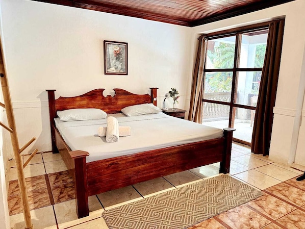 A hotel room with a wooden bed, a window with curtains, a small bedside table with a vase, and a painting on the wall.