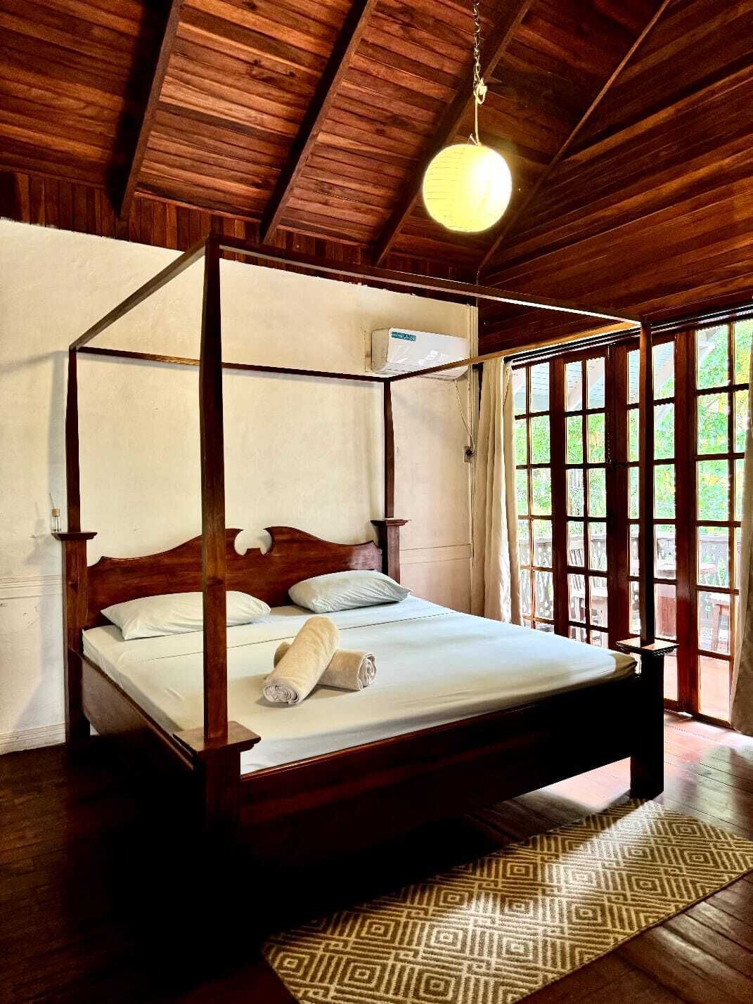 A four-poster bed with a wooden frame and white bedding, a hanging light fixture, and a view of greenery through large windows.