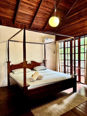 A four-poster bed with a wooden frame and white bedding, a hanging light fixture, and a view of greenery through large windows. - Hotel Mandacaru Puerto Viejo (Cahuita)