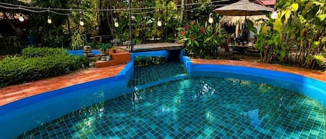 A swimming pool with a tiled edge, surrounded by lush greenery and a thatched-roof structure.