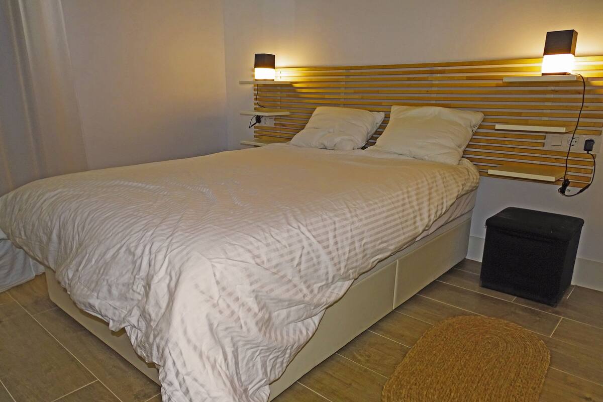 A neatly made bed with a headboard, a bedside table, and a small stool.