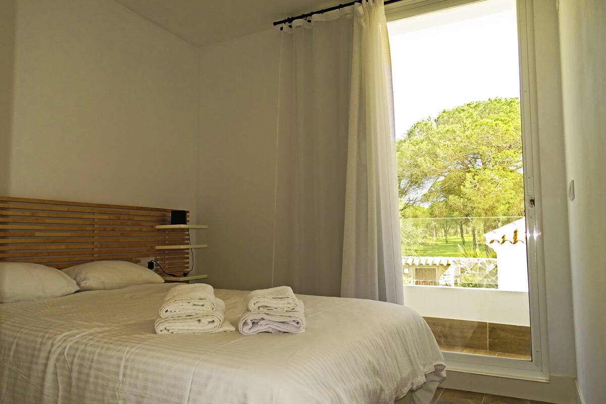 A bedroom with a bed, a wooden headboard, a window with curtains, and a view of greenery outside.