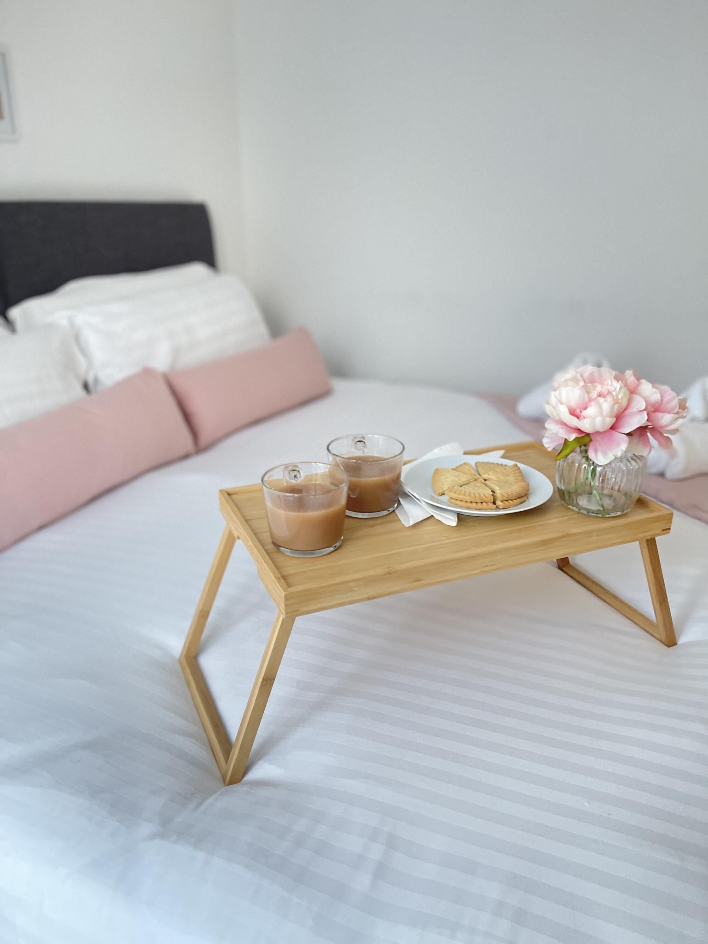 A neatly made bed with a wooden tray holding two glasses of a brown beverage, a plate with cookies, and a vase with flowers.