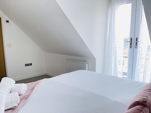 Un dormitorio con techo inclinado, un radiador, una cama con ropa de cama blanca y una manta rosa, y una ventana con cortinas.