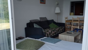 Una habitación con una cama, un sofá, una mesita y una silla. Se ve un ventanal con vistas a zonas verdes.