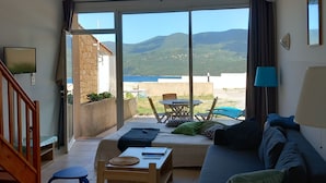 Amplia sala de estar con un gran ventanal corredero que da a un patio exterior. La habitación cuenta con un sofá, una mesa de centro y una mesa de comedor con sillas. Una escalera conduce a un nivel superior.