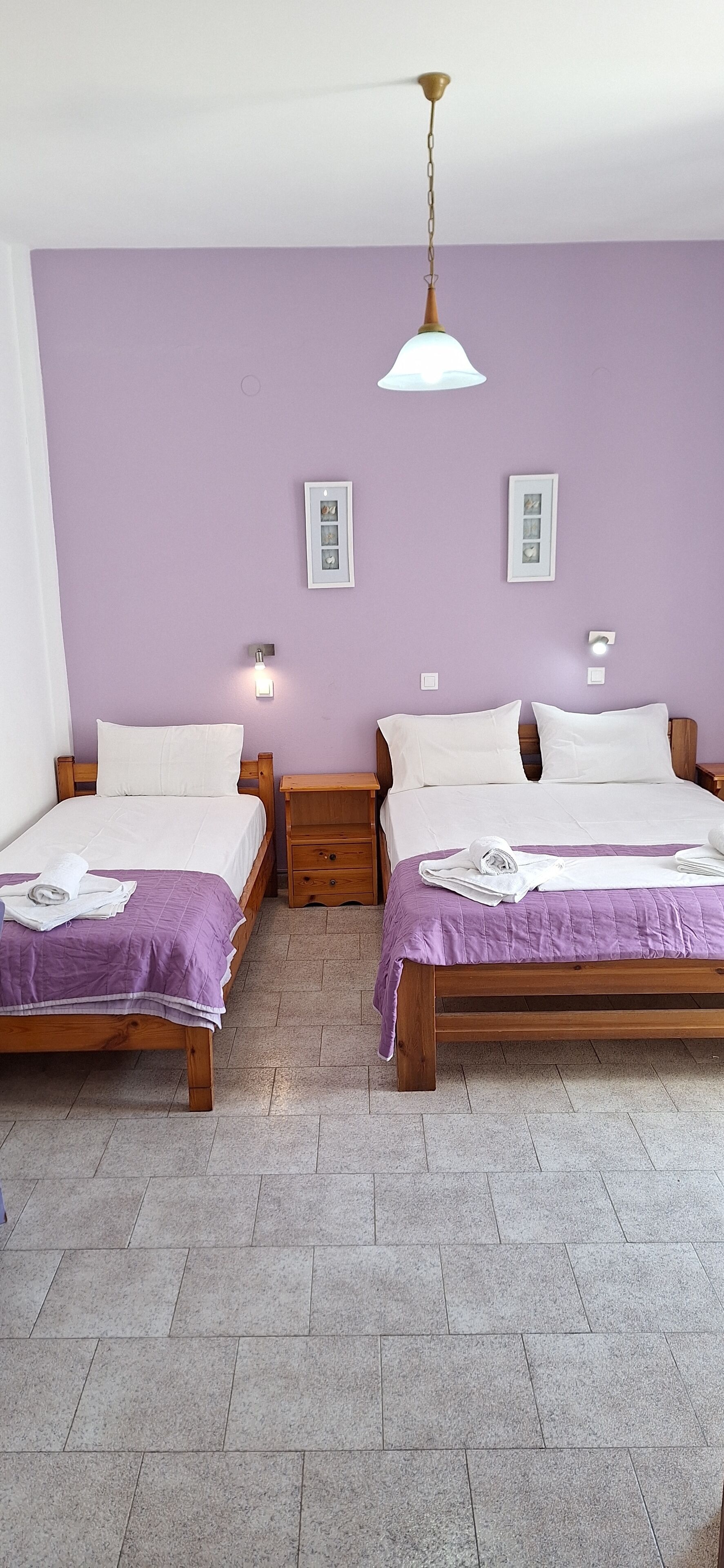Habitación con dos camas, cada una con estructura de madera y ropa de cama morada. Hay mesitas de noche con lámparas e interruptores de luz en la pared.