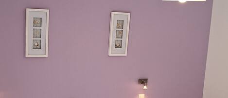 A room with two single beds, each with white linens and purple pillows. There is a wooden nightstand with a lamp between the beds.