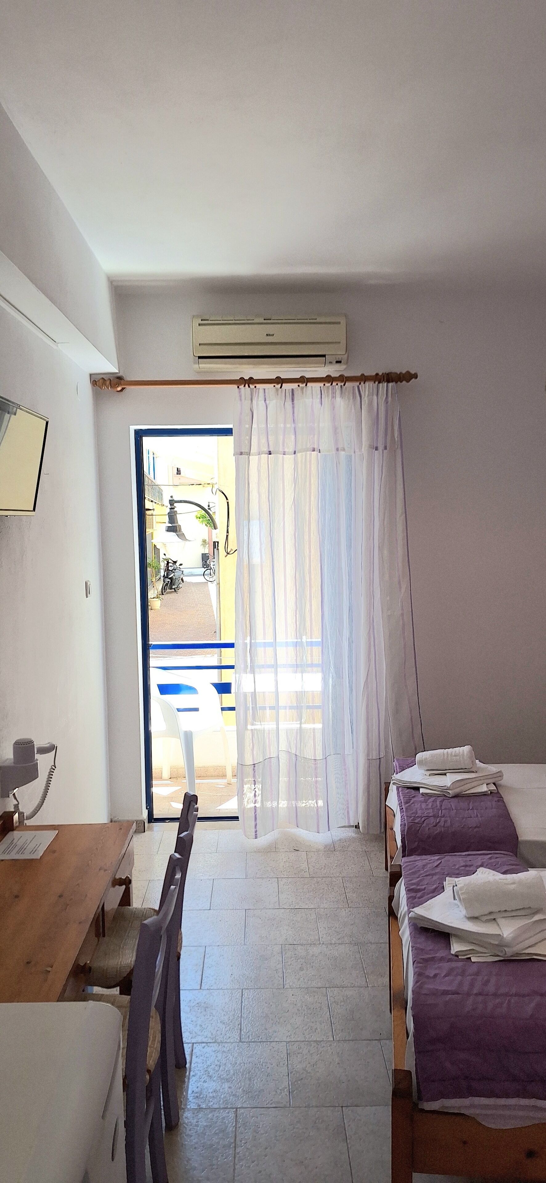 Una habitación con cama, escritorio y silla. Hay una ventana con cortinas, una puerta que da al exterior y un equipo de aire acondicionado en la pared.