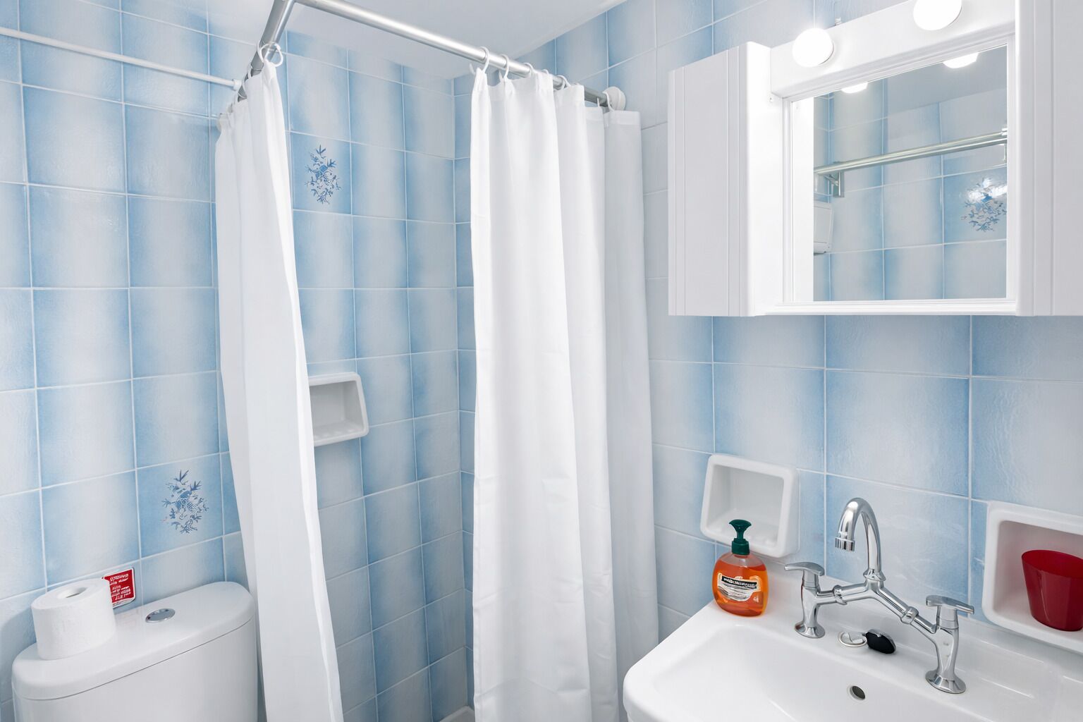 A bathroom with blue tiles, a white shower curtain, a toilet, a sink with a soap dispenser, and a mirror above the sink.