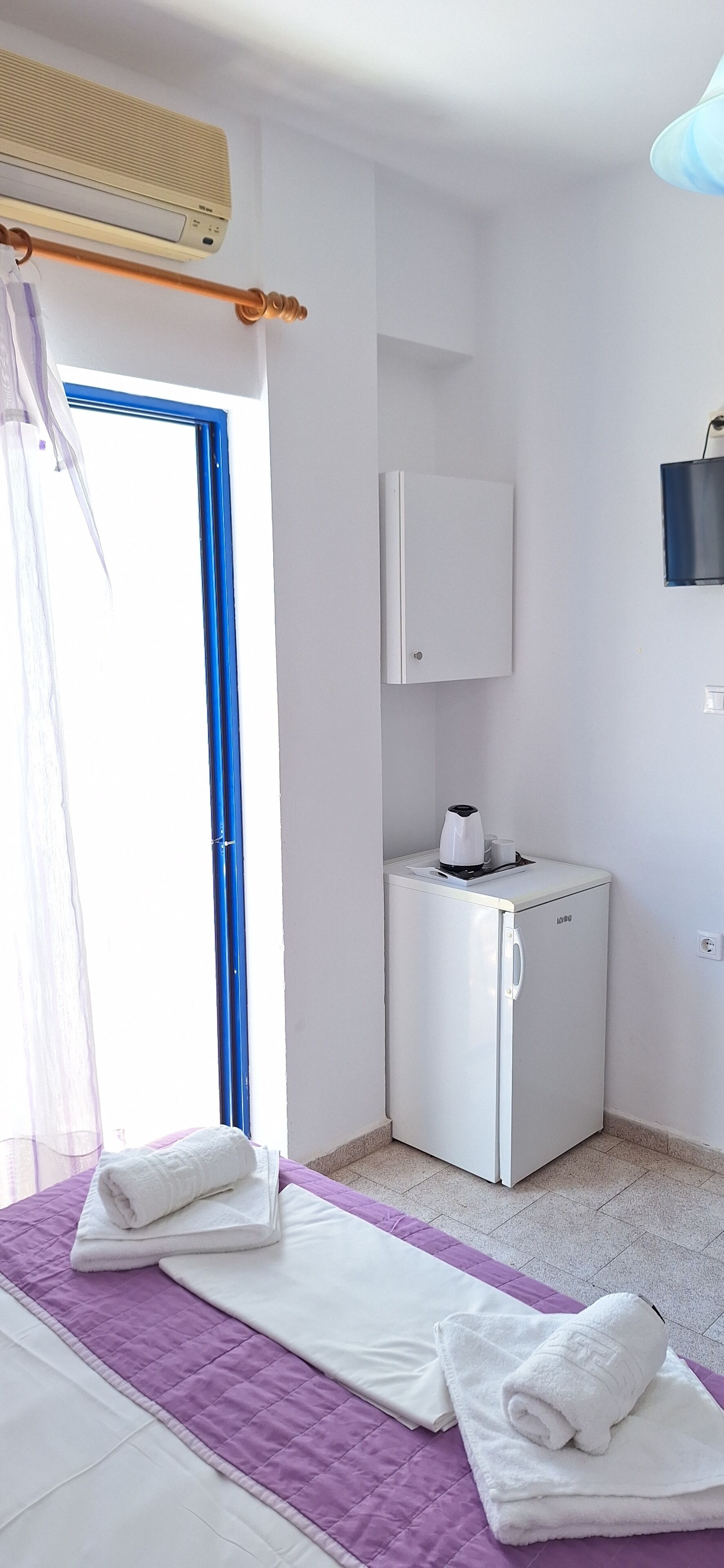 Een kamer met een bed, een kleine witte koelkast, een raam met blauwe lijst en een wandkast.