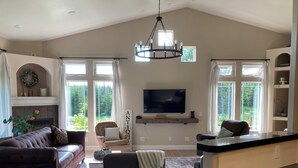 Een ruime woonkamer met een open haard, een televisie en een groot raam met uitzicht op groen.