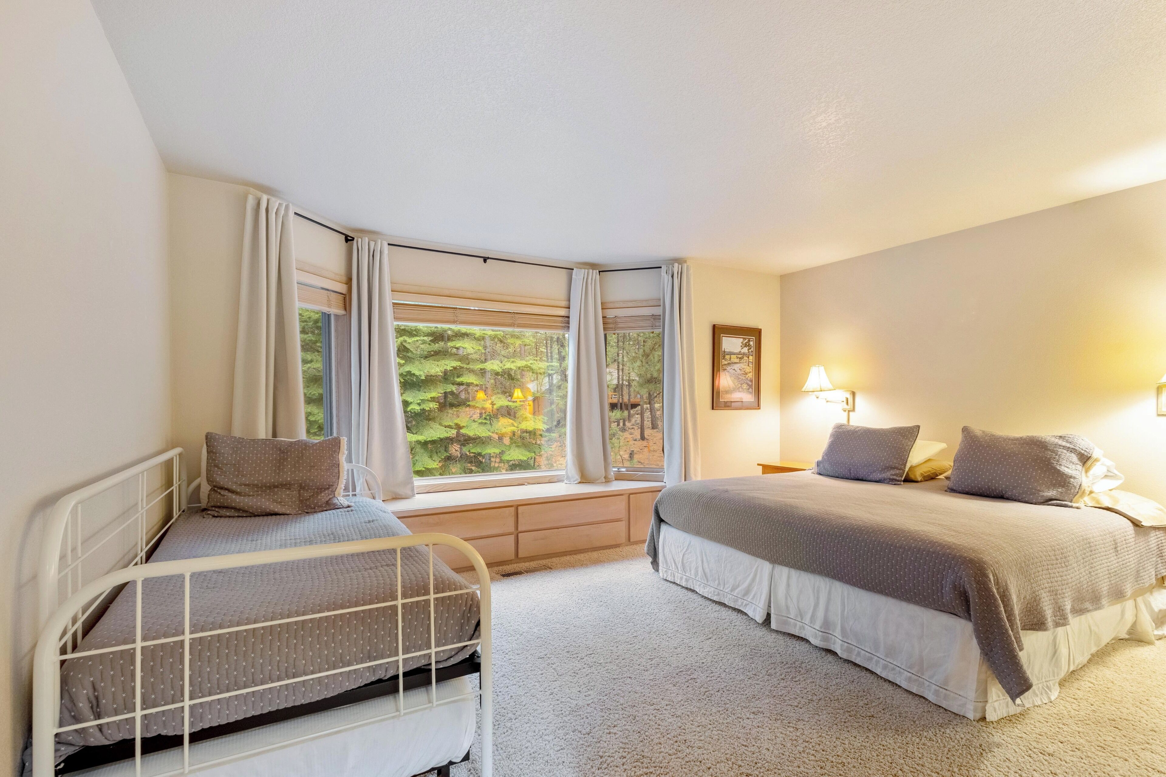 A bedroom with two beds, a window with curtains, a bedside table with a lamp, and a framed picture on the wall.