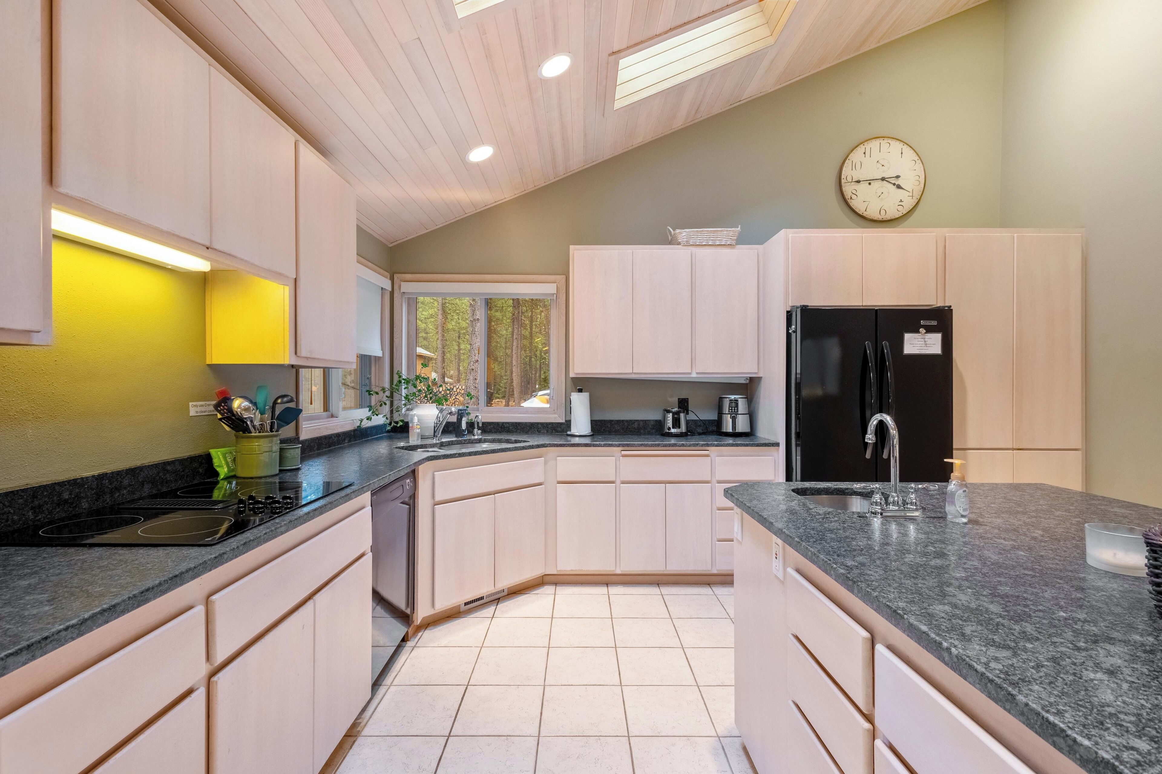 Una cocina moderna con armarios de colores claros, un refrigerador negro y un ventanal con vistas a árboles.