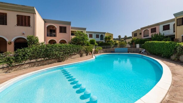 A swimming pool with a clear blue water, surrounded by buildings with arched windows and balconies. - Holiday home (Villasimius)