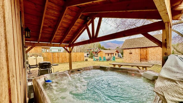 A hot tub area with a wooden roof, a picnic table, and a view of a backyard with houses and mountains. - Stylish Mountain Retreat: Hot Tub, Fire Pit, Whiteface Views! (Wilmington)