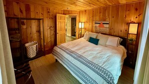 A wooden-paneled bedroom with a bed, a nightstand, a hanging light, and a door leading to another room. - Stylish Mountain Retreat: Hot Tub, Fire Pit, Whiteface Views! (Wilmington)