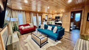 A cozy cabin interior with a red and blue sofa, a glass coffee table, wooden floors, and a kitchen area visible through an open door. - Stylish Mountain Retreat: Hot Tub, Fire Pit, Whiteface Views! (Wilmington)
