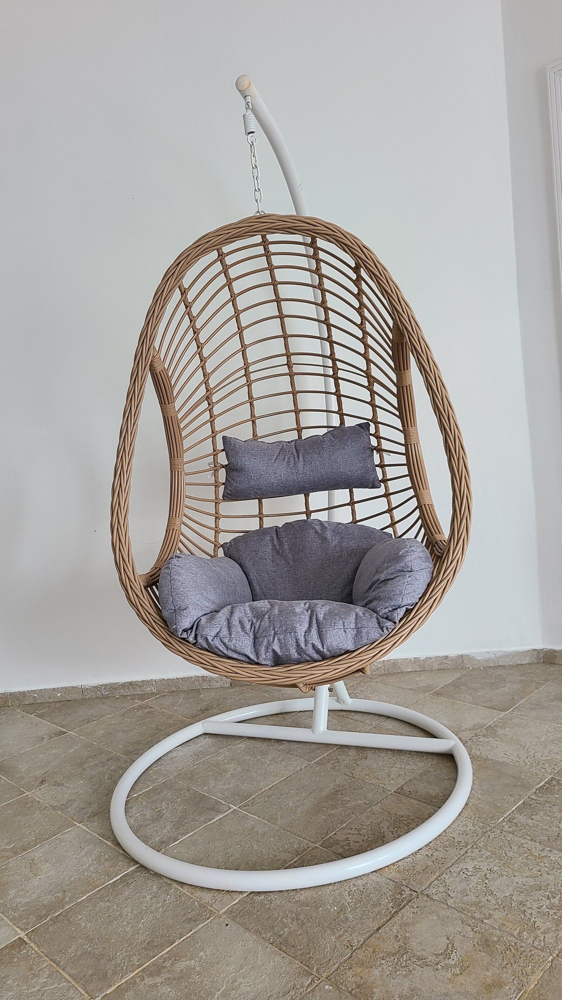 A wicker chair with a cushion and a pillow, placed on a tiled floor against a plain wall.