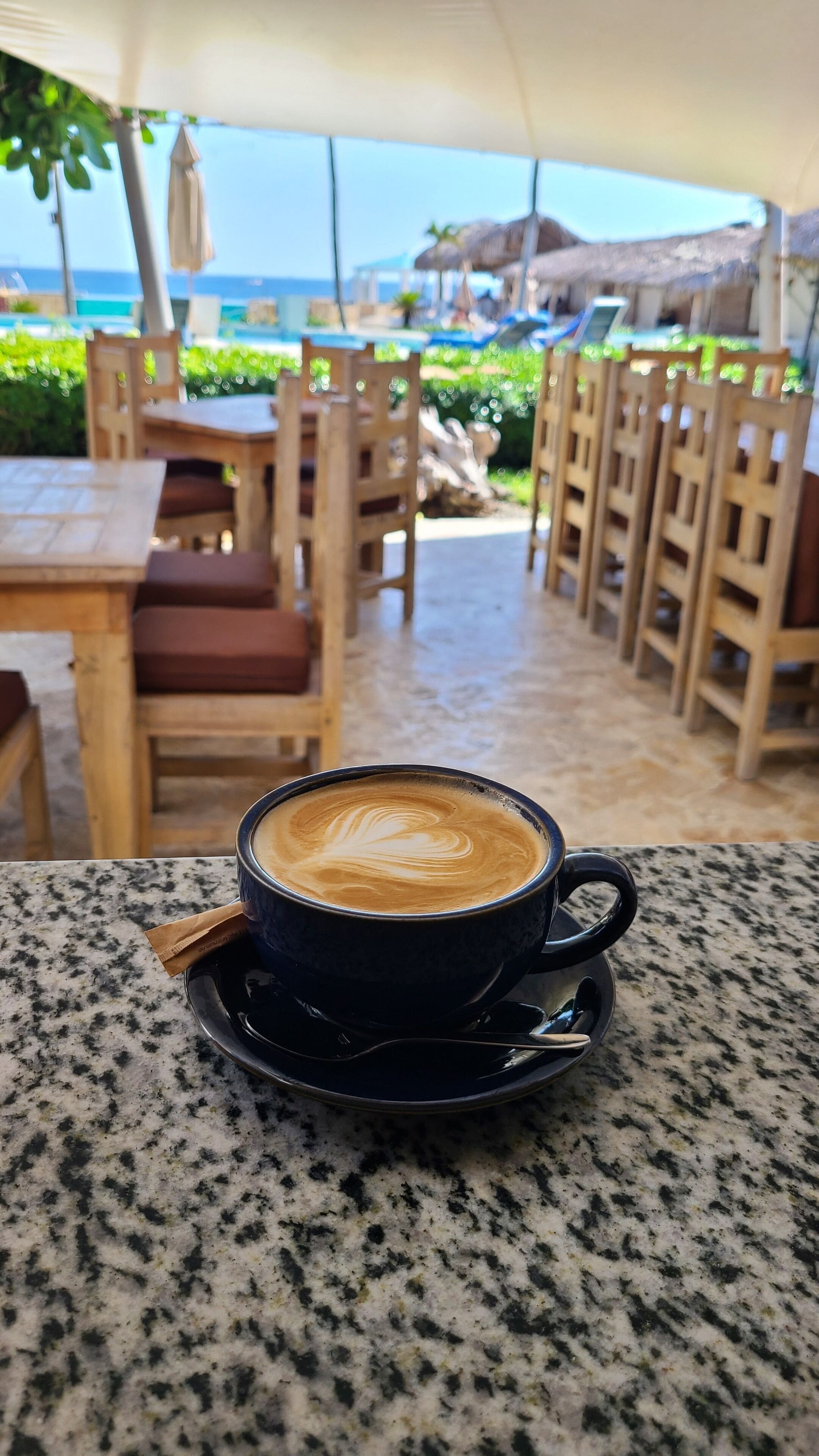 Una taza de café sobre una mesa de granito, con vista a una terraza exterior y una playa al fondo.