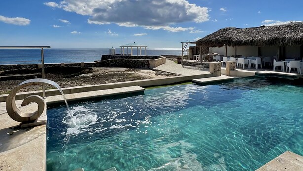Un ciel d’un bleu éclatant surplombe une piscine, une structure à toit de chaume et un belvédère.