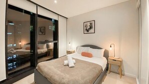 Ein modernes Schlafzimmer mit einem großen Bett, Nachttischen, Blick auf die Stadt durch eine Glastür und einem gerahmten Bild an der Wand.