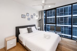 A modern hotel room with a large bed, a nightstand, a desk, a chair, and a large window with a city view.