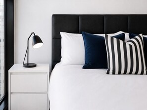 Ein Bett mit weißen und gestreiften Kissen, eine schwarze Lampe auf einem weißen Nachttisch und ein Fenster mit Blick auf Gebäude.