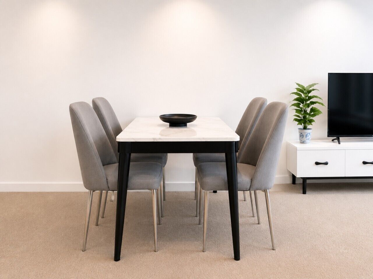 Un coin repas avec une table blanche, quatre chaises grises, un bol noir, un meuble blanc et une plante en pot.