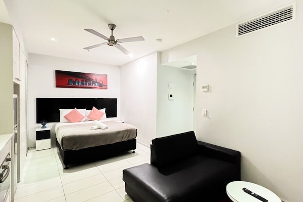 Une chambre d’hôtel avec un lit, un canapé noir, un ventilateur de plafond, un tableau au mur et une table de chevet blanche.