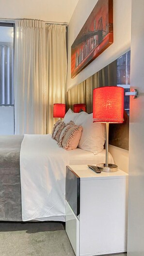 Een moderne hotelkamer met een bed, twee bedlampen, een schilderij en een raam met gordijnen.