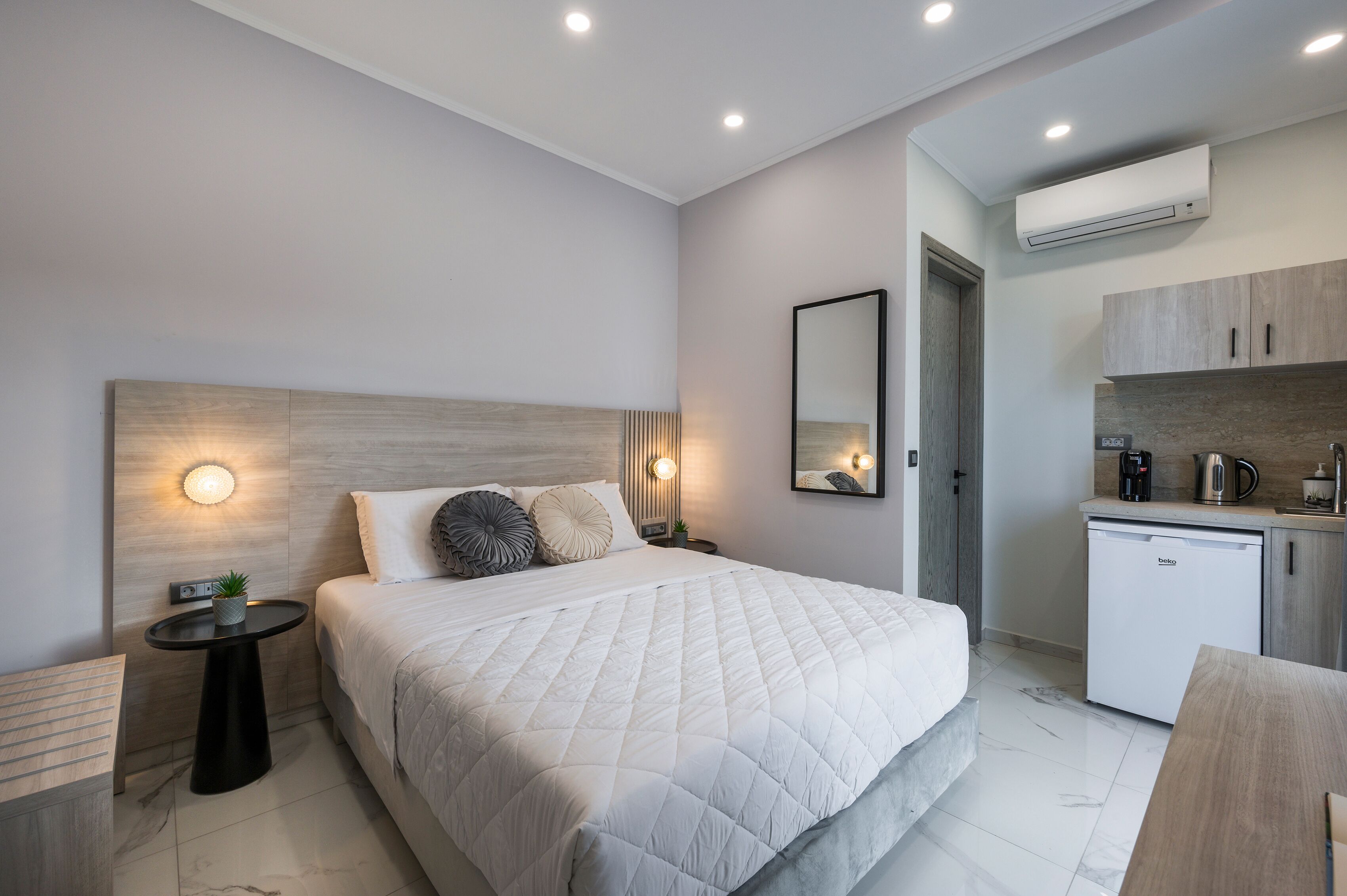 Quarto de hotel moderno com cama grande, mesas de cabeceira, pequena área de cozinha e ar condicionado.