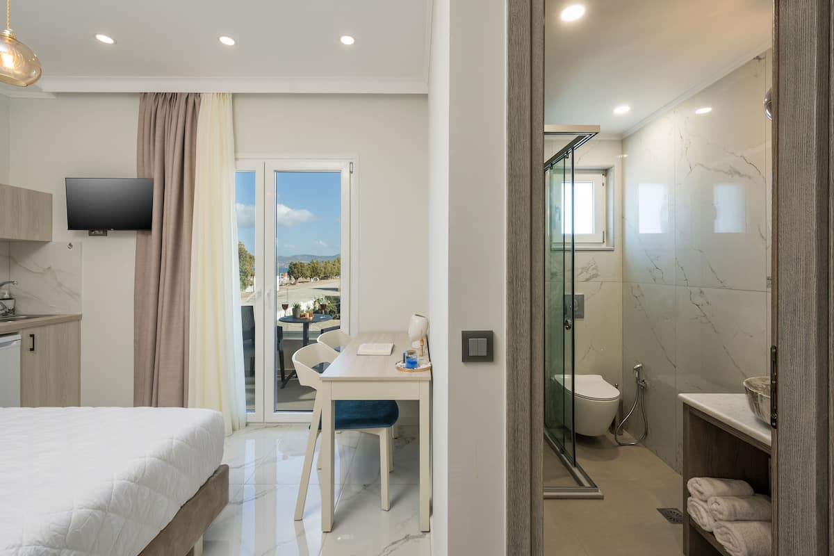 A modern hotel room with a bed, a desk with a chair, a TV, and a bathroom with a glass shower enclosure.