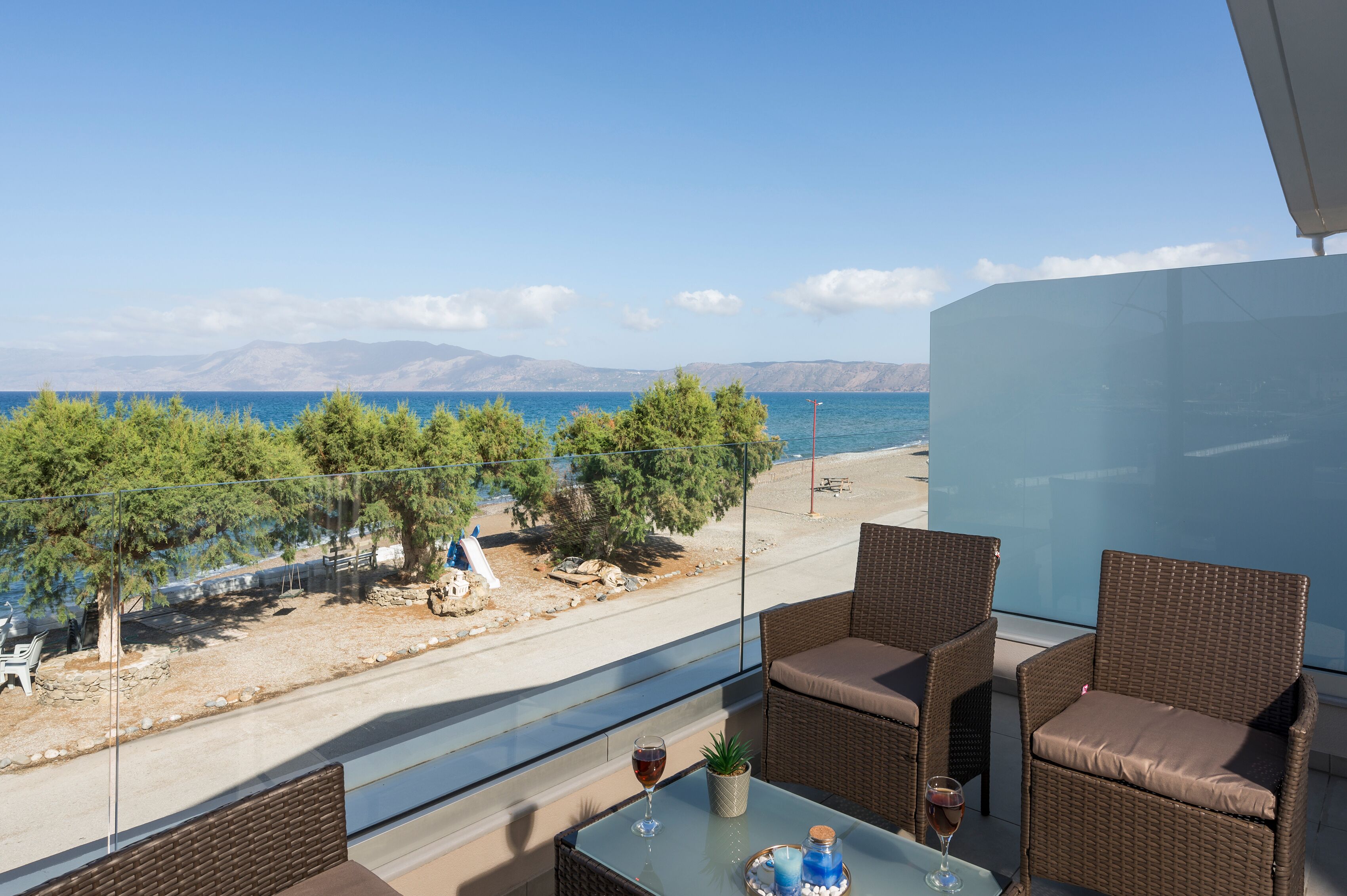A balcony with wicker furniture, a glass table, and a view of the sea and mountains.