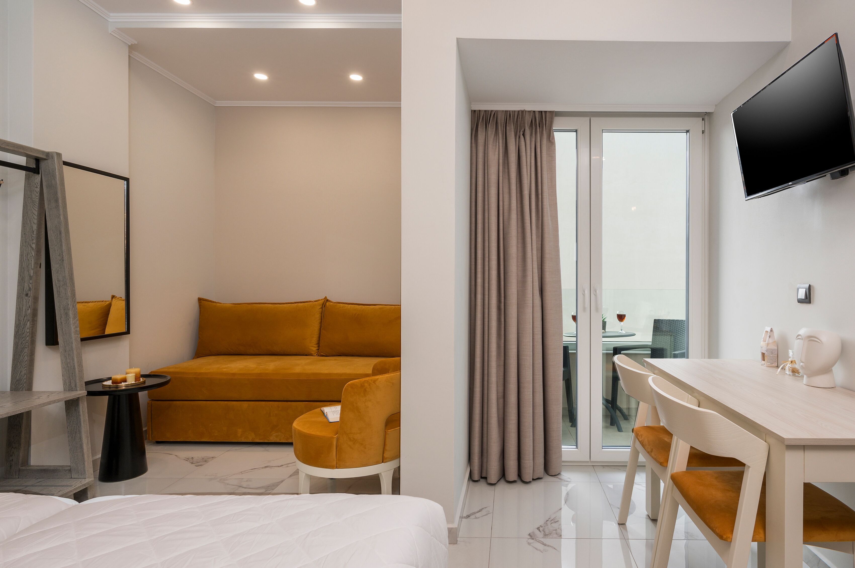 A modern hotel room with a sofa, a small table with candles, a bed, a dining area with a table and chairs, and a TV mounted on the wall.