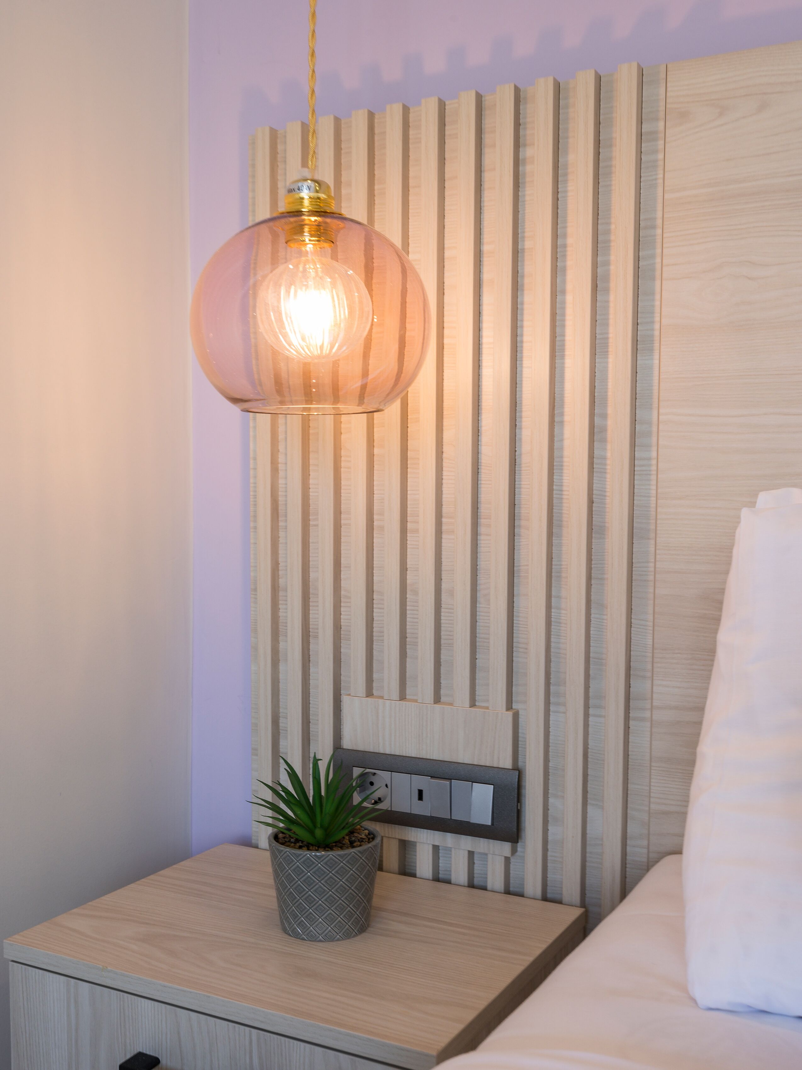 Un dormitorio ordenado con cabecera de madera, una mesita de noche con planta en maceta y una lámpara colgante.