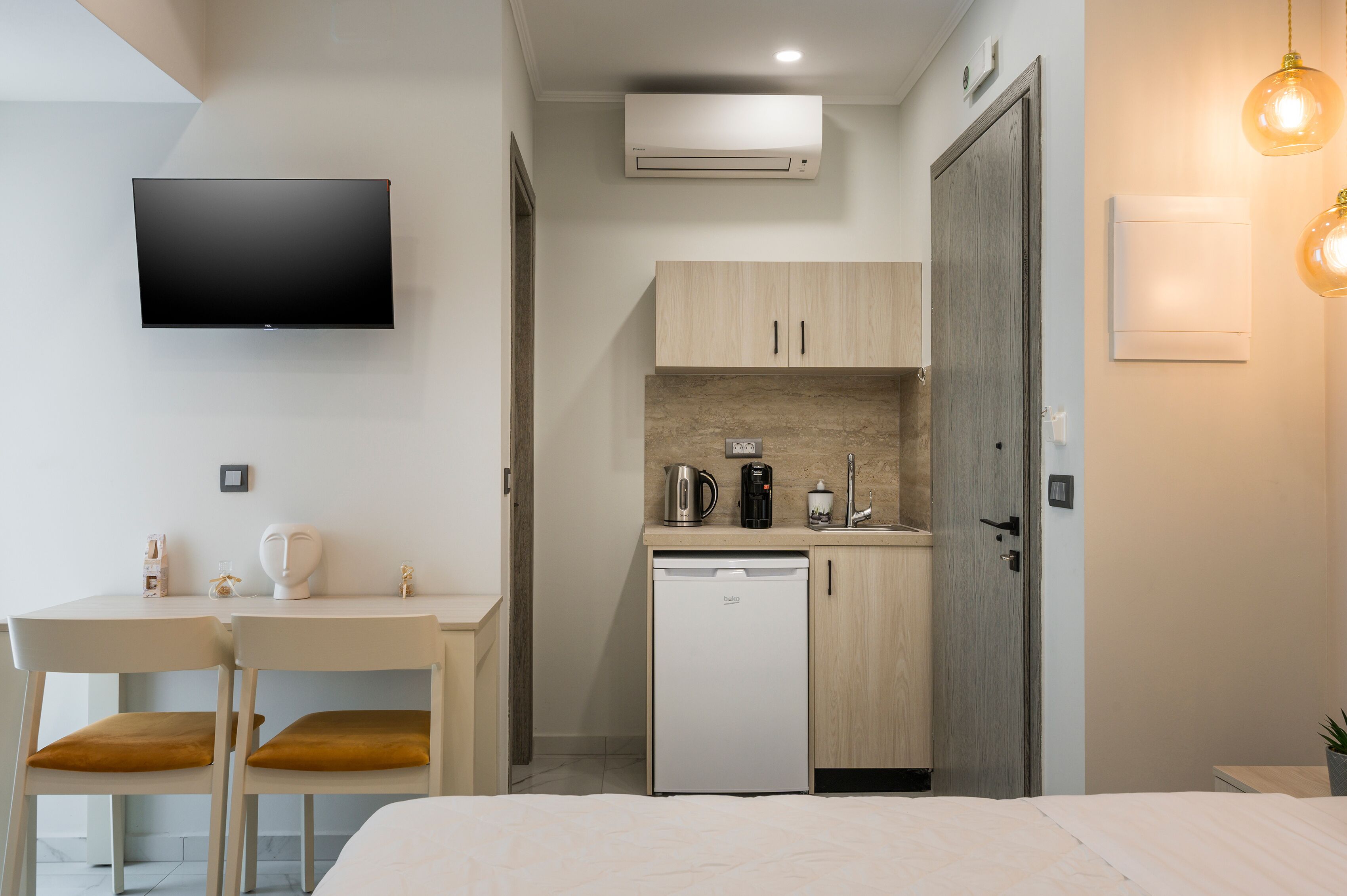Ett kompakt hotellrum med en säng, ett litet pentry med kylskåp, en kaffebryggare och en mikrovågsugn. Det finns en platt-TV monterad på väggen och ett litet bord med stolar.