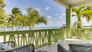 A balcony with wicker chairs and a table, overlooking a beach with palm trees and a thatched-roof structure. - Beach Walkout Condo 112 3BR/2BA Sleeps 8 (Placencia)