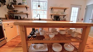 A kitchen with wooden floors, a central island with stacked plates, and various kitchen utensils on shelves. - Le Partage Lodge (Val Morin)