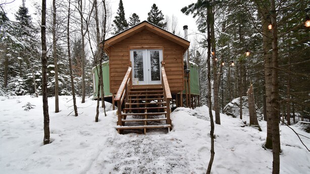 A wooden cabin with a staircase leading to a door, surrounded by snow-covered trees. - Le Partage Lodge (Val Morin)