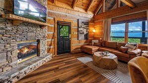 A cozy living room with a stone fireplace, wooden beams, a large TV, and a view of the outdoors.