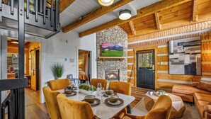 A cozy cabin interior with a stone fireplace, wooden beams, a dining area with a table set for four, and a comfortable seating area.