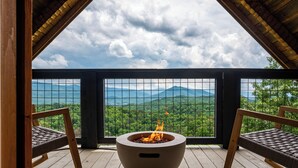 A wooden deck with a fire pit and two wicker chairs, overlooking a mountainous landscape.