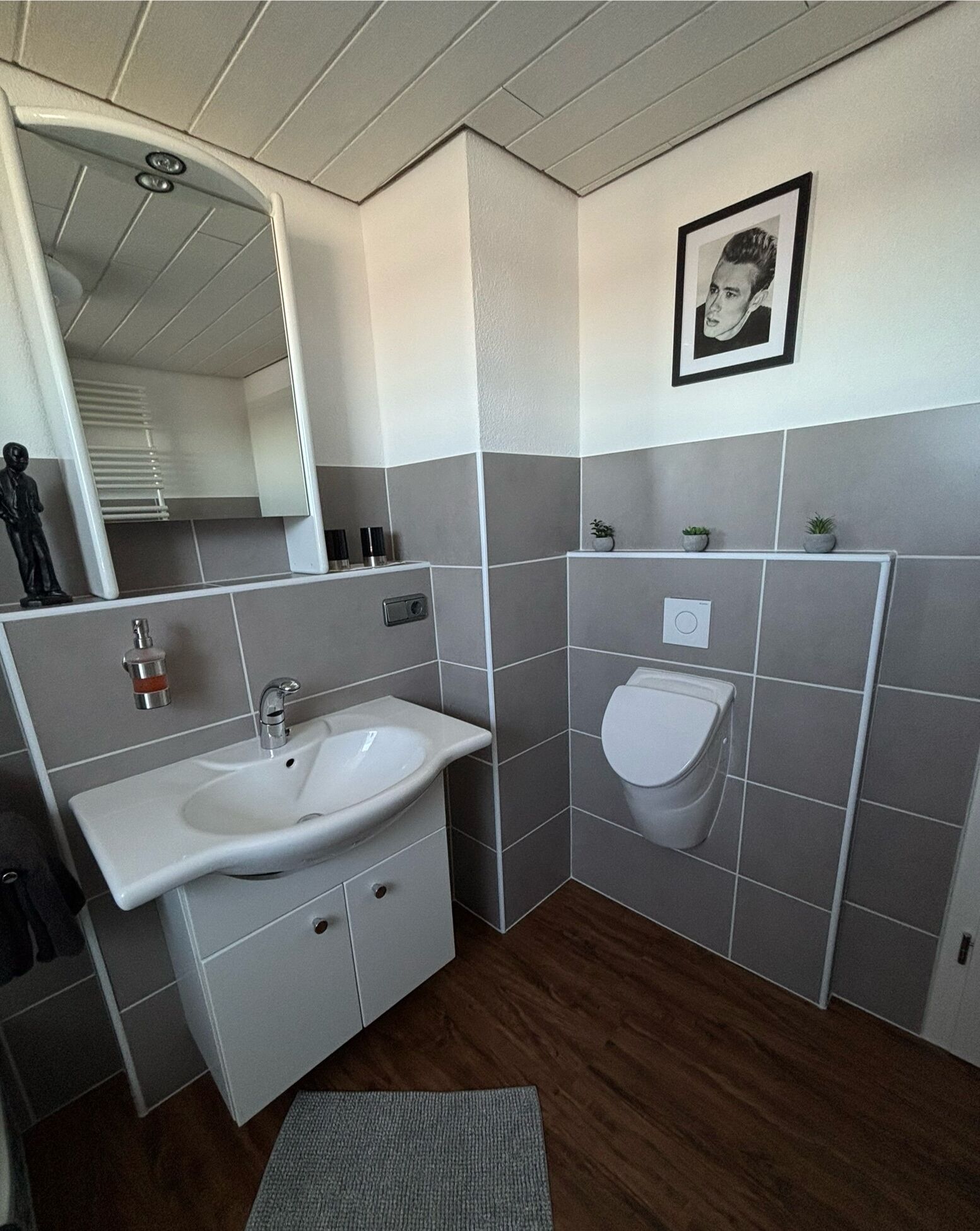 A bathroom with a white sink, a toilet, and a mirror. There are gray tiles on the walls and a wooden floor.