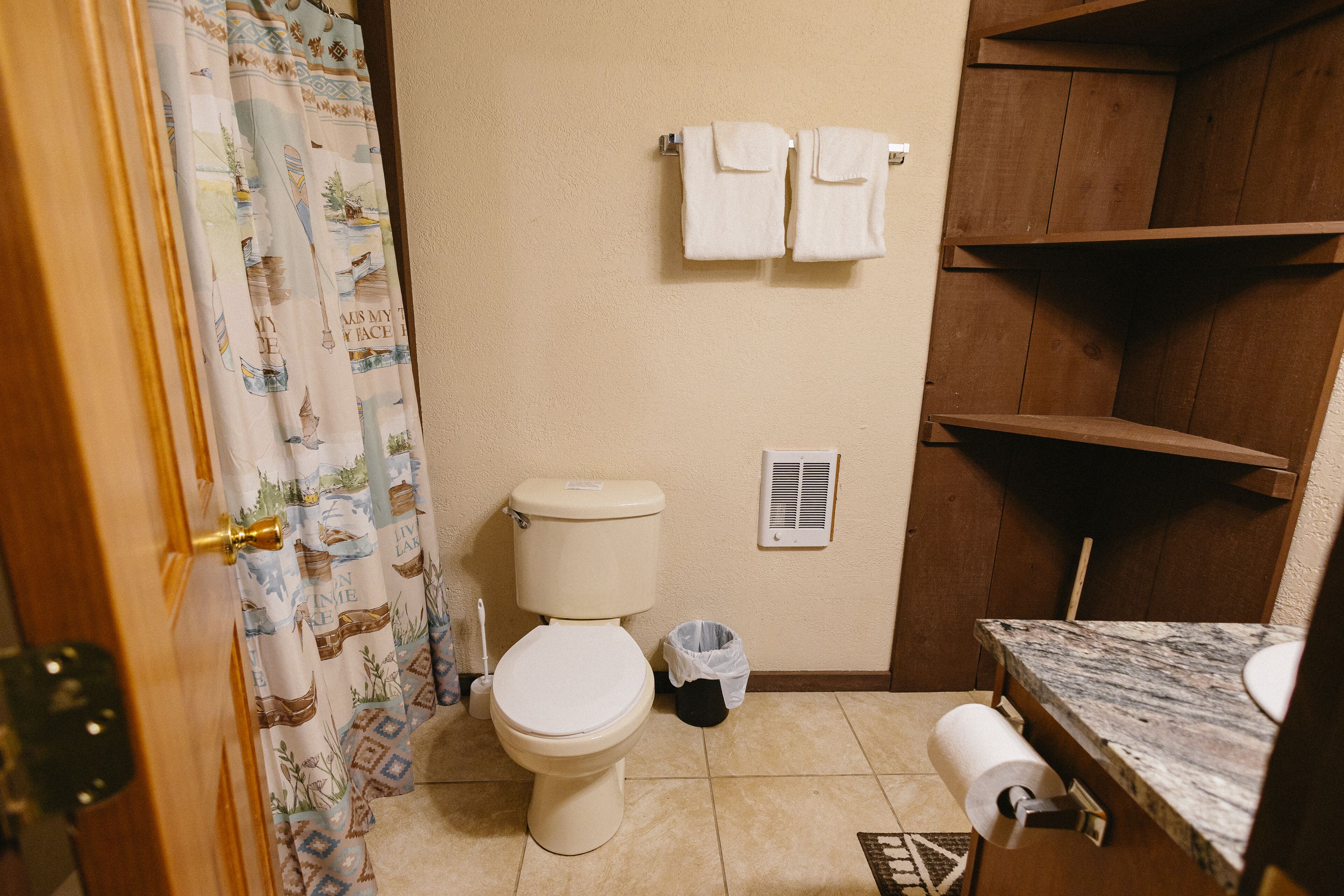 A bathroom with a toilet, a shower curtain, a towel rack with two towels, a trash bin, and a marble countertop with a sink.