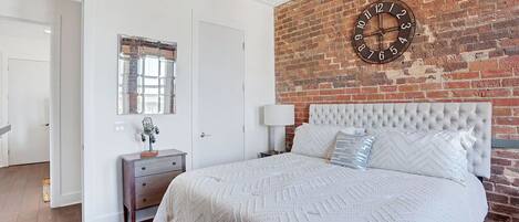 Un dormitorio con pared de ladrillo, un reloj, una cama con cabecera blanca, una mesita de noche y un aparador.