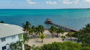 Une plage avec une jetée en bois s’avançant dans des eaux turquoise, bordée de palmiers et d’un bâtiment à la façade verte.